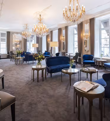 Elegant hotel lounge with opulent chandeliers, large mirrors, and floor-to-ceiling windows. The room features plush blue velvet armchairs and sofas, marble-topped tables, and tall standing lamps, creating a sophisticated atmosphere.