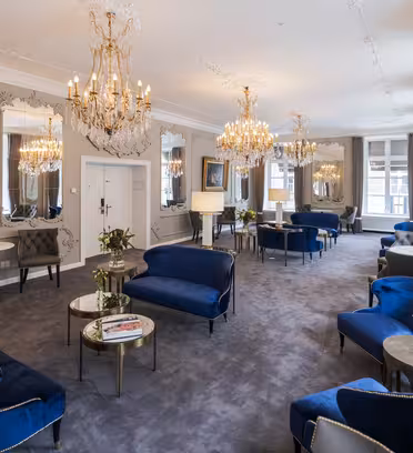 Elegant lounge with plush blue velvet sofas, round tables, and ornate mirrors on the walls. Crystal chandeliers hang from the ceiling, illuminating the room. Large windows with drapes line the side, creating a luxurious and inviting atmosphere.