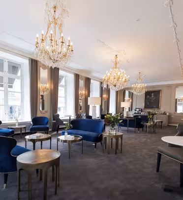 Elegant lounge with velvet blue sofas, gold side tables, and chandeliers. Large windows with gray curtains illuminate the space. Ornate mirrors and decorative molding on walls enhance the classic luxury ambiance.