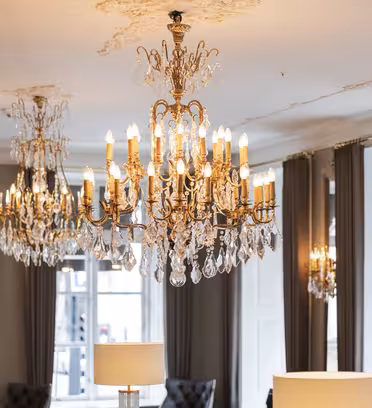 Elegant room with multiple sparkling chandeliers hanging from the ceiling. The room features large windows with white shutters and is decorated with dark drapes and plush chairs, creating a luxurious and sophisticated atmosphere.