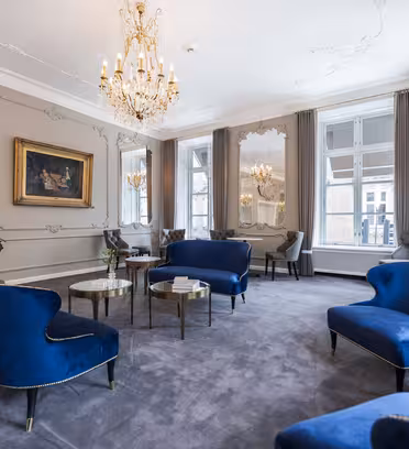 Elegant living room with ornate ceiling, featuring blue velvet chairs and sofas, a chandelier, and large windows with gray curtains. A classic painting hangs on the wall, and round glass tables are arranged throughout the space.