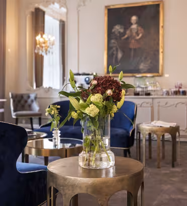 Elegant lounge with blue velvet chairs, a round table with a floral vase centerpiece featuring red and white flowers. Large windows and an ornate chandelier enhance the sophisticated ambiance. Artwork and refreshments in the background add a cozy touch.