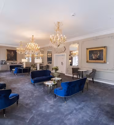 Elegant lounge with blue velvet chairs, round tables, and ornate chandeliers. The room features large mirrors, a plush gray carpet, and artwork on the walls. Classical and luxurious ambiance with natural light streaming through windows.