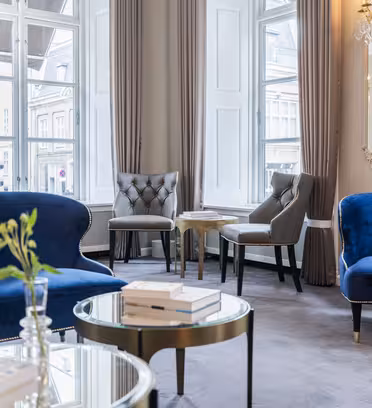 Elegant living room with large windows, blue velvet sofas, gray upholstered chairs, and round glass coffee tables. Soft lighting from a chandelier reflects off ornate mirrors, creating a cozy, sophisticated atmosphere.