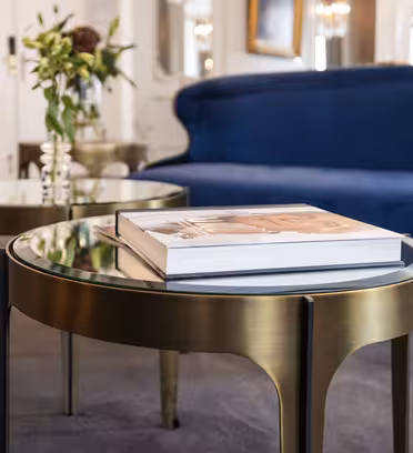 A modern living room features a sleek brass coffee table with a glass top, holding a book. In the background, a deep blue velvet sofa and a side table with a vase and flowers add elegance to the neutral-toned carpeted space.