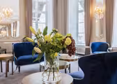 A luxurious room with blue velvet chairs, a glass vase with green and red flowers, and small round tables. Large windows and elegant chandeliers add to the sophisticated ambiance. The walls are adorned with mirrors and intricate molding.