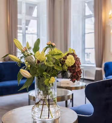 A luxurious room with blue velvet chairs, a glass vase with green and red flowers, and small round tables. Large windows and elegant chandeliers add to the sophisticated ambiance. The walls are adorned with mirrors and intricate molding.