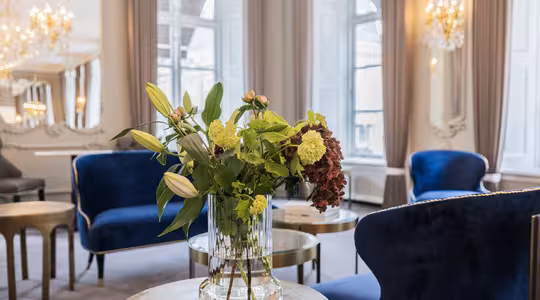 A luxurious room with blue velvet chairs, a glass vase with green and red flowers, and small round tables. Large windows and elegant chandeliers add to the sophisticated ambiance. The walls are adorned with mirrors and intricate molding.