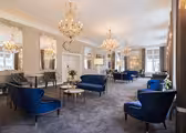 An elegant, spacious room with blue velvet seating, large ornate mirrors, and several golden chandeliers hanging from the ceiling. The walls are a soft neutral tone, and there are flower arrangements on small round tables.