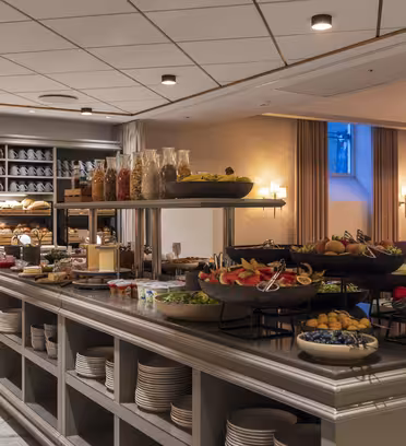 A buffet-style hotel dining area with a variety of food options on display, including fruits, vegetables, cereals, and baked goods. The room is warmly lit, with elegant shelving and neatly arranged dishes on the counter.