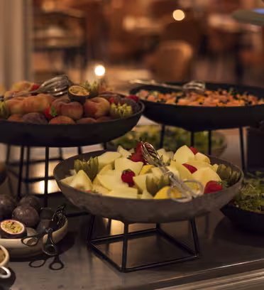 A buffet table with a variety of fresh fruits and salads. Black bowls hold sliced apples, pears, kiwis, peaches, and passion fruits. Tongs rest in some of the bowls, and the background shows a blurred dining area.
