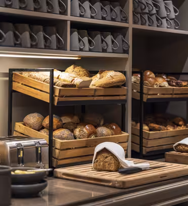 A bakery display with wooden crates holding assorted breads, including loaves and rolls. A cutting board with sliced bread and a bread knife is on the counter. Shelves above have stacked cups and tea boxes. The scene is warmly lit.