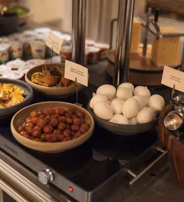 A buffet table with a selection of food. Includes a bowl of hard-boiled eggs, a dish of meatballs, a bowl of tortilla chips, cheese on a stand, and a basket with condiments. Yogurt containers are in the background.