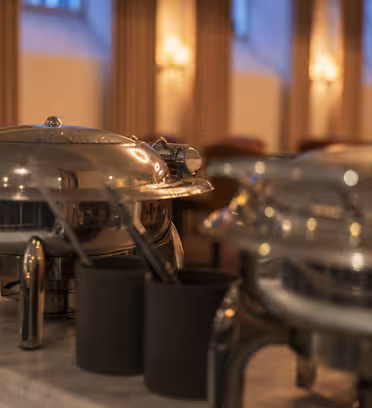 Two shiny metallic chafing dishes on a marble counter, each with a lid and handle. Black utensil holders sit nearby. The background features blurred warm lighting and soft drapery in a dimly lit room.