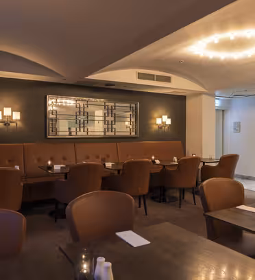 A dimly lit restaurant interior with brown leather chairs and dark wooden tables. A cushioned bench lines the wall beneath modern wall sconces and a decorative mirror. The room is softly illuminated by a circular ceiling light.