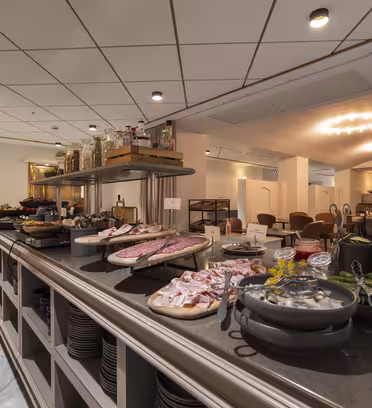 A restaurant buffet featuring various dishes including meats, vegetables, and other appetizers. The setting is elegant with neatly placed tables and ambient lighting, creating a welcoming atmosphere.