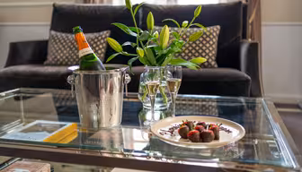 A glass table holds a plate of chocolate-covered strawberries, two filled champagne glasses, and a champagne bottle in an ice bucket, with a vase of lilies and a sofa in the background.