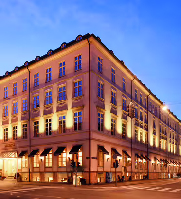 A grand, illuminated building at dusk with rows of windows and architectural details. Awning-covered entrances are visible at the street level. The sky is a rich blue, and streetlights cast a warm glow on the empty intersection.