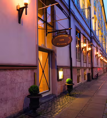 Street view of a restaurant, Restaurant von Plessen, with warm lighting on the buildings exterior. The sidewalk is lined with small green plants in pots. A bicycle is parked further down the pathway.
