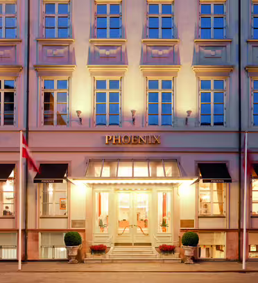The image shows the facade of a building named Phoenix. It features multiple evenly spaced windows, three flagpoles with flags, and a lit entrance with steps leading up. Potted plants flank the doorway, giving a welcoming appearance.