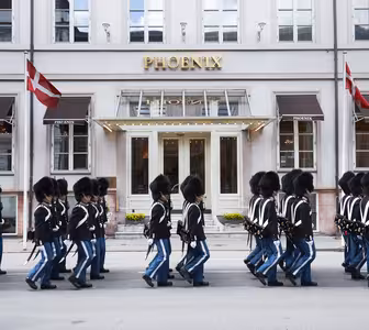 A line of Royal Danish Guards in traditional uniform march past the Hotel Phoenix in Copenhagen. The guards wear black hats and blue pants. Danish flags are displayed on the building.