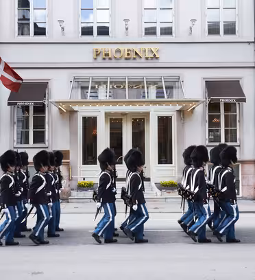 A line of Royal Danish Guards in traditional uniform march past the Hotel Phoenix in Copenhagen. The guards wear black hats and blue pants. Danish flags are displayed on the building.