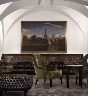 A stylish lounge with patterned armchairs and a green velvet chair centered on a carpet. A large painting of a scenic village landscape hangs on a curved, white wall, illuminated by wall sconces. A round ottoman with a geometric pattern is in the foreground.