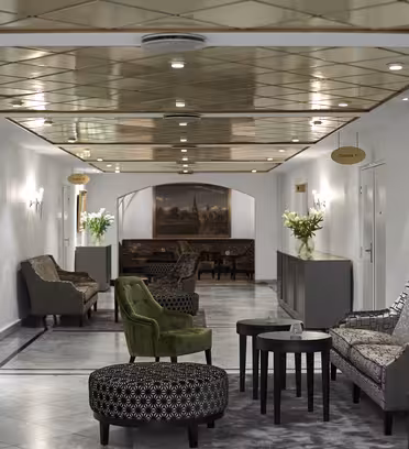 A sophisticated hotel hallway with elegant armchairs, patterned ottomans, and small tables lining the marble floor. Artistic paintings and soft lighting create a luxurious atmosphere, complemented by fresh flower arrangements on the tables.