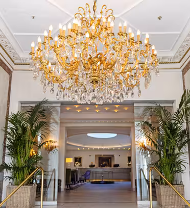 Elegant hotel lobby with a large, ornate chandelier hanging from a decorative ceiling. Marble walls and grand mirrors enhance the space. Potted plants flank the entrance to a reception area with soft lighting and modern seating.