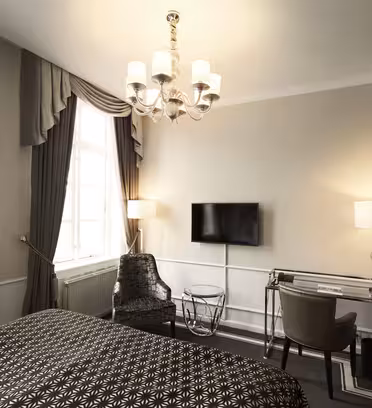A hotel room with a double bed covered in a patterned duvet. Theres a window with curtains, a wall-mounted TV, and a seating area with a chair and glass-topped table. A desk with a chair is on the right, under a suspended lamp and next to a minibar.
