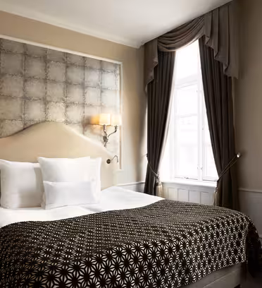 Luxurious hotel room with a large bed adorned with patterned brown and white bedding. Soft lighting from wall sconces flanks a textured silver headboard. Elegant drapes frame a tall window, allowing natural light to illuminate the space.