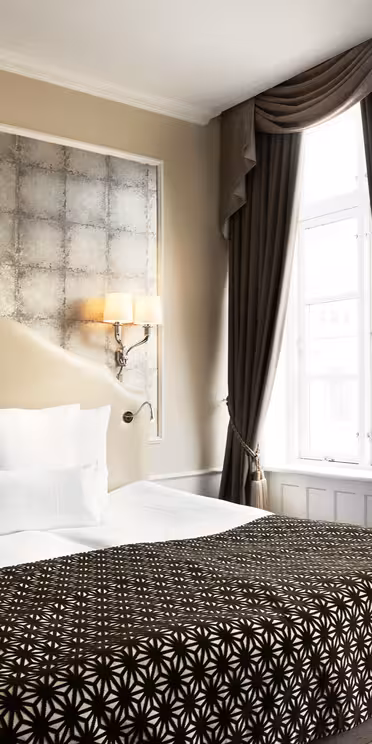 Luxurious hotel room with a large bed adorned with patterned brown and white bedding. Soft lighting from wall sconces flanks a textured silver headboard. Elegant drapes frame a tall window, allowing natural light to illuminate the space.