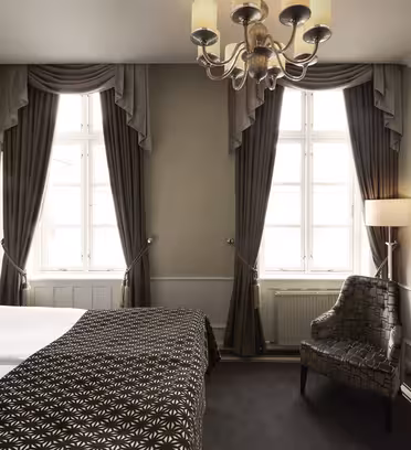 Hotel room with a neatly made bed with patterned bedding, two tall windows with dark drapes, a patterned armchair, a wall-mounted TV, and a chandelier. The room is softly lit, and the walls are painted in neutral tones.