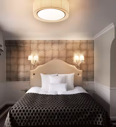 A cozy hotel room features a neatly made bed with a dark patterned blanket. Two lit sconces flank a textured headboard. Light filters through a window with draped curtains on the left. A ceiling fixture softly illuminates the room.