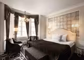 Elegant hotel room with a large bed, accented by a patterned headboard and decorative pillows. Two chairs and a small table are near the window, covered by long curtains. A modern chandelier and wall lamps provide lighting, with a patterned carpet underfoot.