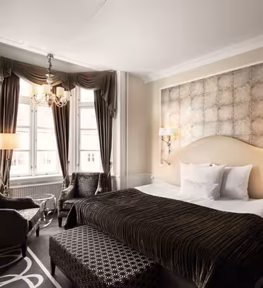 Elegant hotel room with a large bed, accented by a patterned headboard and decorative pillows. Two chairs and a small table are near the window, covered by long curtains. A modern chandelier and wall lamps provide lighting, with a patterned carpet underfoot.
