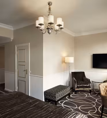 A modern hotel room with a large wall-mounted TV, elegant chandelier, patterned carpet, and a mix of armchairs and a bench. Neutral-toned walls and lighting give the space a cozy feel.