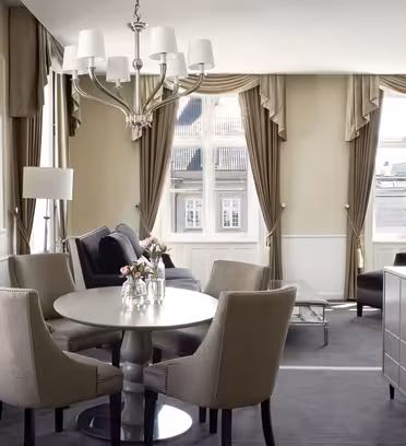 A well-lit, elegant hotel room with beige and white decor. It features a round table with four upholstered chairs, a black sofa, a chandelier, and large windows with beige curtains, offering a city view.