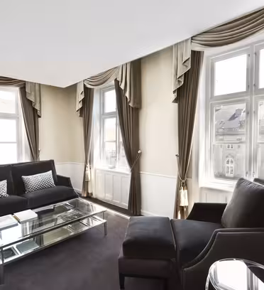 A sophisticated living room with large windows, brown and beige curtains, and dark upholstered furniture. A glass coffee table sits between two chairs and a sofa. Sunlight filters through the windows, highlighting the elegant decor.