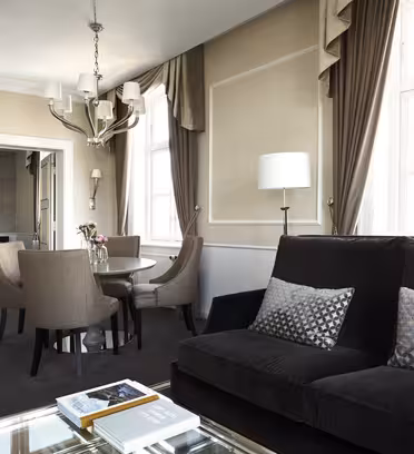 Elegant hotel suite featuring a sitting area with a dark sofa, patterned pillows, and a glass coffee table. A round dining table with beige chairs is nearby. The adjoining bedroom is visible, fitted with a bed and decorative chandelier. Large windows brighten the space.