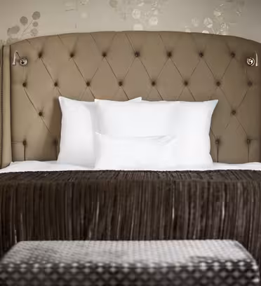 A neatly made bed with a tufted headboard, flanked by two wall-mounted lamps. The bedding features white pillows and a brown textured throw. Two bedside tables are visible, set against a wall with subtle floral-patterned wallpaper.