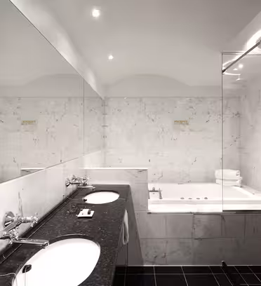 A modern bathroom with marble walls features a large mirror, double sinks with a black countertop, and a built-in Jacuzzi tub. The lighting is soft, highlighting the clean and minimalist design. Towels are neatly folded by the tub.