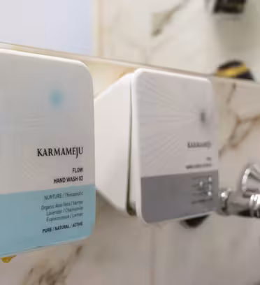 Close-up of two Karmameju hand wash dispensers mounted on a marble wall near a shiny faucet. The dispensers have a modern design and are labeled FLOW HAND WASH 02 and NURTURE Therapeutic, with blue and gray color accents.