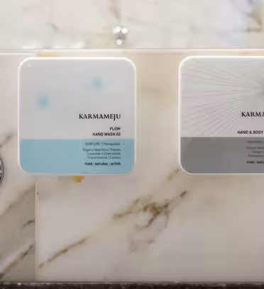 Two square Karmameju dispensers are mounted on a marble wall above a metal sink fixture. The left dispenser is labeled Flow, and the right is labeled Chill. Both feature minimalist designs with soft colors.