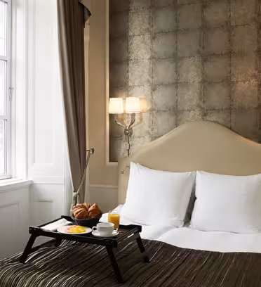 Elegant hotel room with a cozy bed, featuring white pillows and a dark bedspread. A breakfast tray with croissants, orange juice, and fruit is on the bed. The room has a textured accent wall, soft lighting, and a window with brown curtains.
