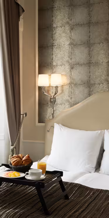 Elegant hotel room with a cozy bed, featuring white pillows and a dark bedspread. A breakfast tray with croissants, orange juice, and fruit is on the bed. The room has a textured accent wall, soft lighting, and a window with brown curtains.
