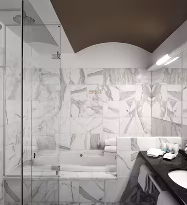 Modern bathroom with marble walls and floor, a glass-enclosed shower, and a tub. Theres a dark countertop with a sink, toiletries, and towels neatly arranged. An overhead fluorescent light illuminates the space.
