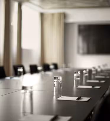 A long, empty conference room table with black chairs is set with clear glasses and pads of paper with pens. A large, blurred screen or board is visible on the wall at the far end. The room is softly lit by natural light from windows.