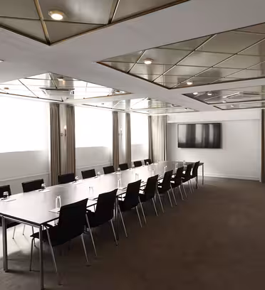 A modern conference room with a long table surrounded by black chairs. The room features a mirrored ceiling with geometric patterns and has beige curtains on the windows. A large flat-screen TV is mounted on the wall.