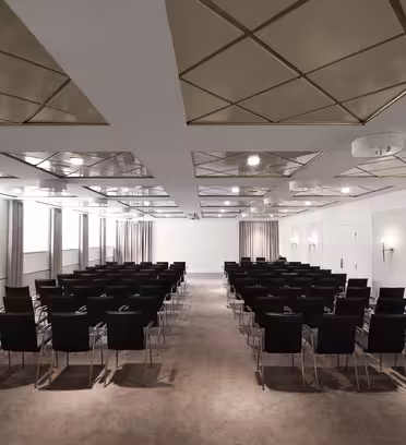 A modern conference room with rows of black chairs facing forwards. The ceiling features geometric gold panels and lights. The walls are white, with minimal decorations. Curtains cover some windows, and the floor is carpeted in a neutral tone.